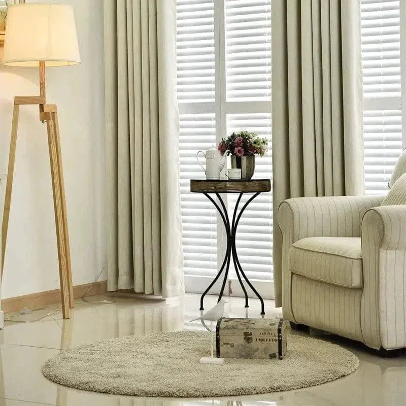 Salon élégant avec un fauteuil confortable et un tapis rond beige, illuminé par une lampe sur trépied. Table d'appoint noire avec décorations florales, devant une fenêtre avec des rideaux et des stores. Ambiance lumineuse et moderne, idéale pour un intérieur contemporain.