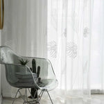 Chaise design transparente placée devant des rideaux blancs à motif végétal, créée pour embellir un intérieur moderne avec élégance. Présence d'une décoration minimaliste comprenant un cactus et des livres, ajoutant une touche contemporaine à l'ensemble. Ambiance lumineuse et épurée idéale pour un salon raffiné.
