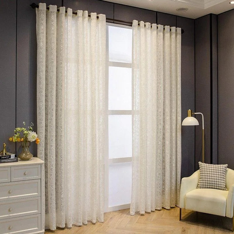 Rideaux élégants ornés de motifs délicats suspendus dans un salon moderne, offrant un éclairage tamisé et créant une ambiance chaleureuse. La pièce est aménagée avec une chaise rembourrée et une lampe au design contemporain, accentuée par des fleurs fraîches sur une commode.