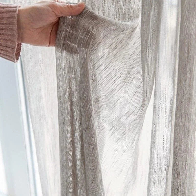 Rideau translucide beige laissant passer la lumière, idéal pour une ambiance lumineuse et moderne dans le salon ou la chambre, main tenant le tissu pour en montrer la fluidité et la finition soignée.
