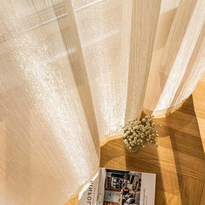 Rideau lumineux offrant une ambiance apaisante avec lumière naturelle filtrée, posé sur un sol en bois chaleureux. Décoration intérieure sophistiquée avec une touche de nature, grâce à un bouquet de fleurs blanches délicatement disposé. Livre ouvert sur des idées de design moderne.