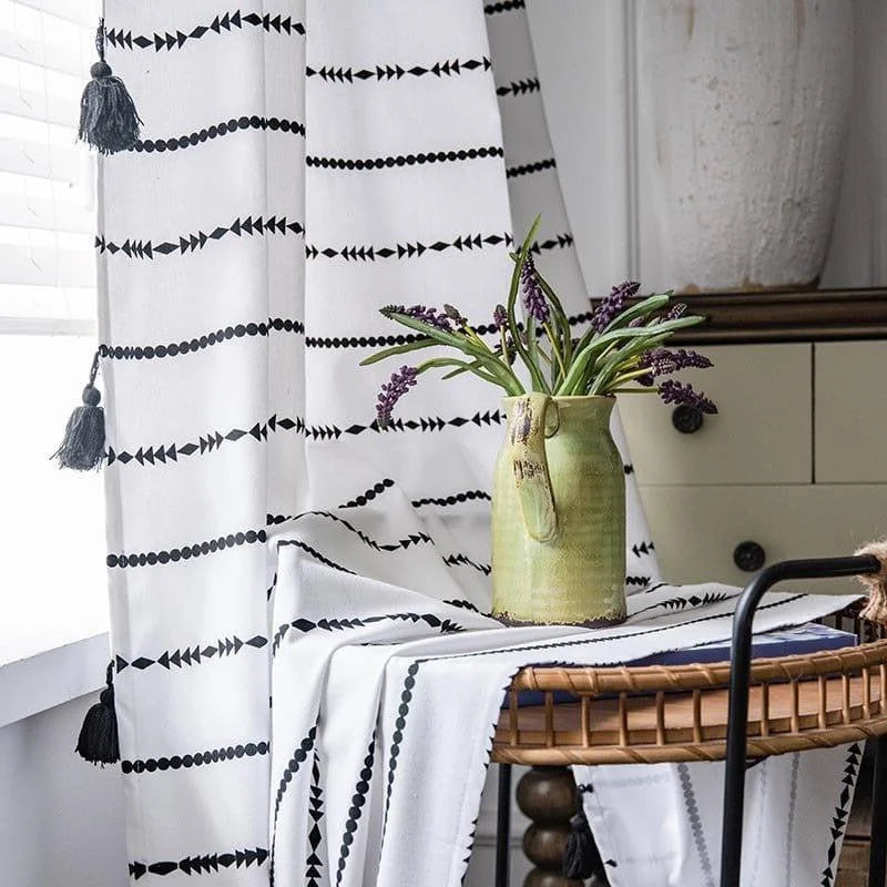 Rideau blanc avec motifs géométriques noirs, suspendu à une fenêtre lumineuse, accompagné d'un vase vert contenant des fleurs violettes, apportant une touche élégante à la décoration intérieure.