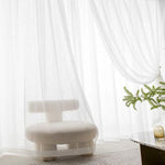 Fauteuil design moderne blanc dans un salon lumineux avec rideaux élégants et plante verte sur une table basse. Ambiance minimaliste et épurée idéale pour un intérieur contemporain et raffiné.