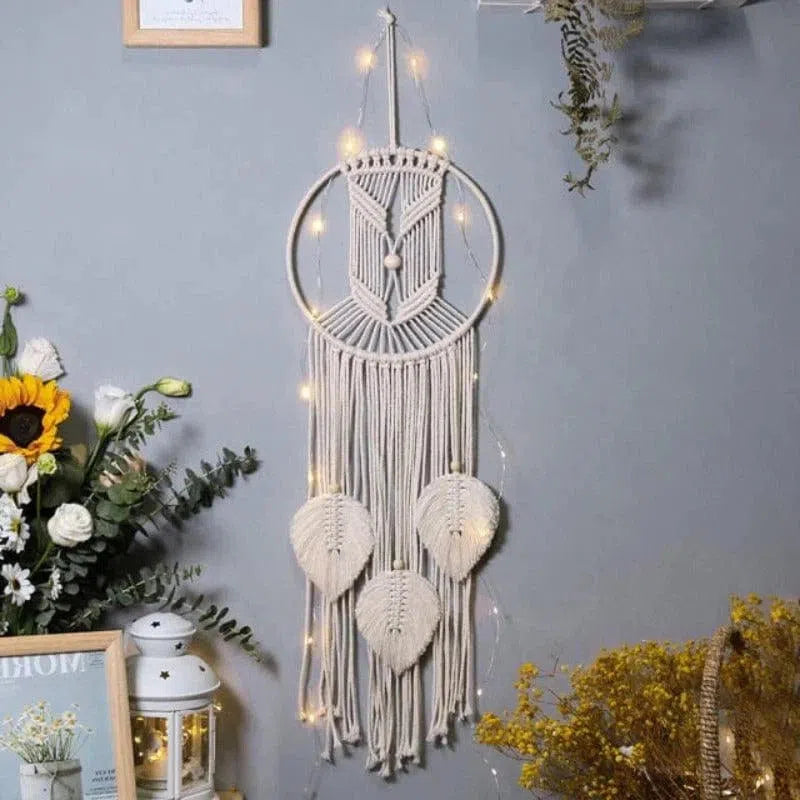 Décoration murale bohème attrape-rêves avec motifs de feuilles, illuminée par des guirlandes lumineuses, sur fond de mur gris, entourée de plantes décoratives et fleurs colorées. Parfait pour une ambiance intérieure chaleureuse et apaisante.