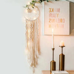 Attrape-rêves lumineux avec guirlande suspendu au mur, créant une atmosphère bohème et chaleureuse. Décoration murale élégante à côté de bougies dorées sur un support, ajoutant une touche de douceur et de tranquillité à votre intérieur. Parfait pour un salon ou une chambre à coucher.