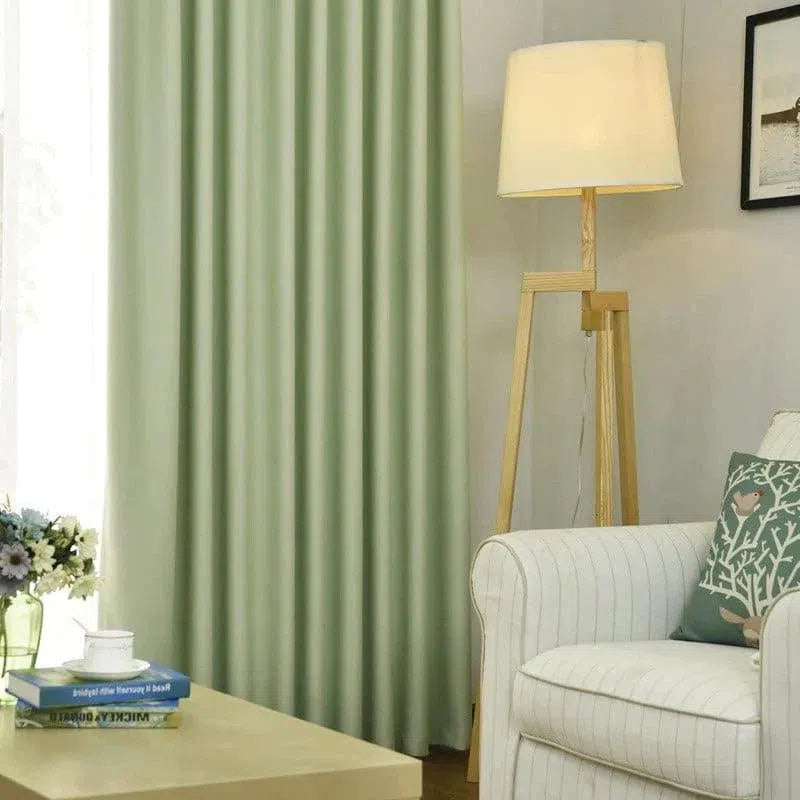 Salon moderne avec rideaux verts, lampe sur pied en bois et fauteuil blanc à rayures fines. Décoration intérieure élégante avec des coussins décoratifs sur le canapé, une table basse avec livres et fleurs en vase. Atmosphère accueillante et lumineuse.