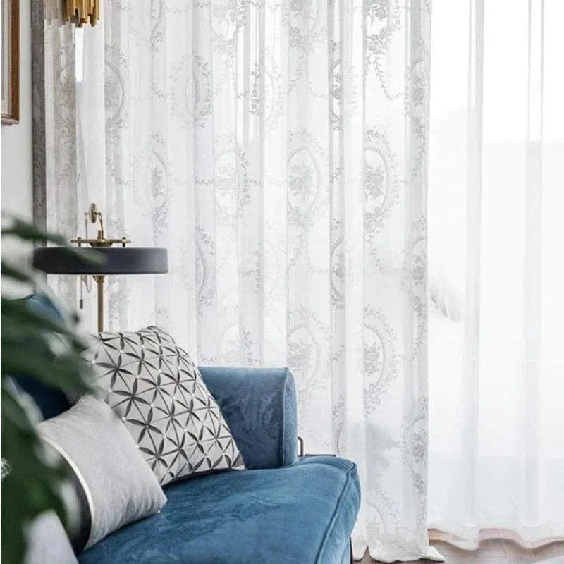 Rideaux élégants à motif floral translucide laissant passer la lumière, parfaits pour une ambiance lumineuse et raffinée dans un salon moderne. Accentués par un canapé bleu et un coussin à motifs géométriques, ces rideaux apportent une touche de charme et de douceur à la décoration intérieure.