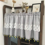 Petite brise-bise décorative grise avec motifs brodés et franges colorées à pompons jaunes et bleus, idée élégante pour habiller une étagère ou une petite fenêtre, ajoutant une touche de charme et de modernité à votre intérieur.