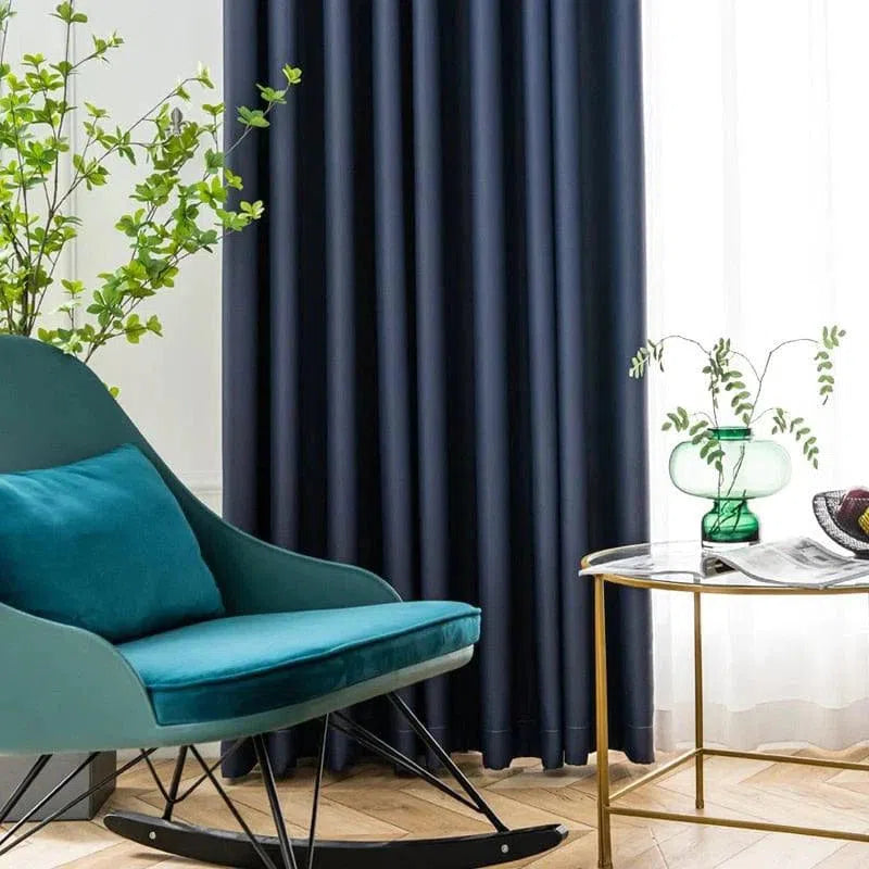 Fauteuil à bascule design en bleu avec coussin assorti, situé dans un salon moderne. Les rideaux sombres ajoutent une touche d'élégance, tandis que la table dorée avec vase vert apporte une touche de fraîcheur. Un espace idéal pour se détendre et lire dans un cadre harmonieux et stylé.