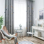 Chambre lumineuse et moderne avec rideaux gris à motifs géométriques, apportant une touche élégante et contemporaine. Espace aménagé avec fauteuil confortable, petite table d'appoint et chaise design, idéal pour un coin lecture ou détente. Ambiance chaleureuse avec décoration minimaliste et parquets en bois clair.