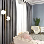 Rideaux opaques gris élégants dans un salon moderne, combinés avec un mobilier chic comprenant un canapé beige aux accents cloutés, un fauteuil doré avec un coussin moelleux et une lampe originale à deux globes lumineux. Un tapis texturé et une plante verte en coin ajoutent une touche naturelle et chaleureuse à la décoration intérieure.