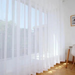 Rideau blanc translucide suspendu dans un salon lumineux avec vue sur l'extérieur, apportant une ambiance douce et aérée à la pièce, idéal pour la décoration intérieure moderne.