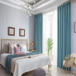 Chambre élégante avec rideaux bleu clair et lit double, décorée de coussins multicolores et d'un jeté de lit, illuminée par un lustre moderne. Ambiance lumineuse et apaisante grâce à la grande fenêtre encadrée de rideaux. Mobilier assorti avec une chaise design et des accents dorés, créant un espace harmonieux et accueillant pour le repos.