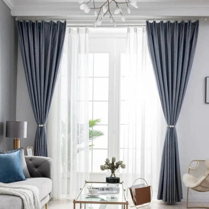 Rideaux opaques bleu foncé élégamment disposés dans un salon moderne, accompagnés de voilages blancs transparents. Le décor présente un canapé gris avec des coussins bleus, une table basse en verre, et un éclairage en suspension qui ajoute une touche de sophistication. Idéal pour une ambiance chaleureuse et contemporaine dans votre espace de vie.