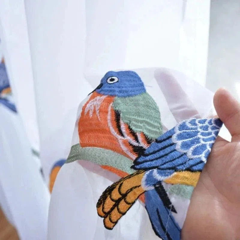 Image d'un motif délicatement brodé représentant un oiseau coloré sur un fond clair. Le design vibrant inclut des nuances de bleu, orange et vert, offrant une touche artistique et élégante pour la décoration intérieure. Parfait pour apporter une ambiance naturelle et chaleureuse à votre espace de vie. Idéal pour les amateurs de motifs animaliers et de décoration unique.