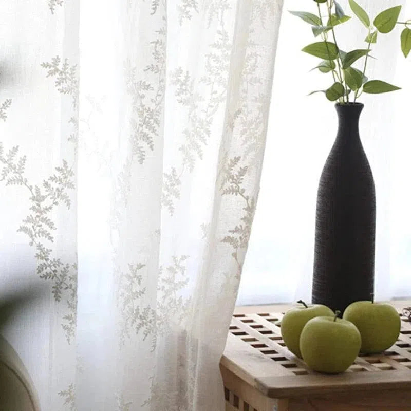 Rideau translucide élégant avec motif floral, laissant passer une douce lumière naturelle. Décoration moderne avec vase noir et pommes vertes sur une table en bois, créant une ambiance apaisante et raffinée pour la maison.