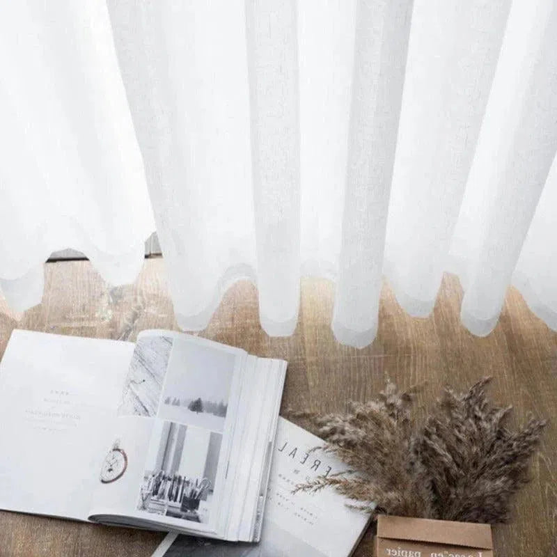 Voilage blanc élégant laissant passer la lumière naturelle, idéal pour un salon moderne. Photographie d'un ouvrage à pages ouvertes sur parquet clair avec un bouquet décoratif. Parfait pour une décoration intérieure lumineuse et épurée.