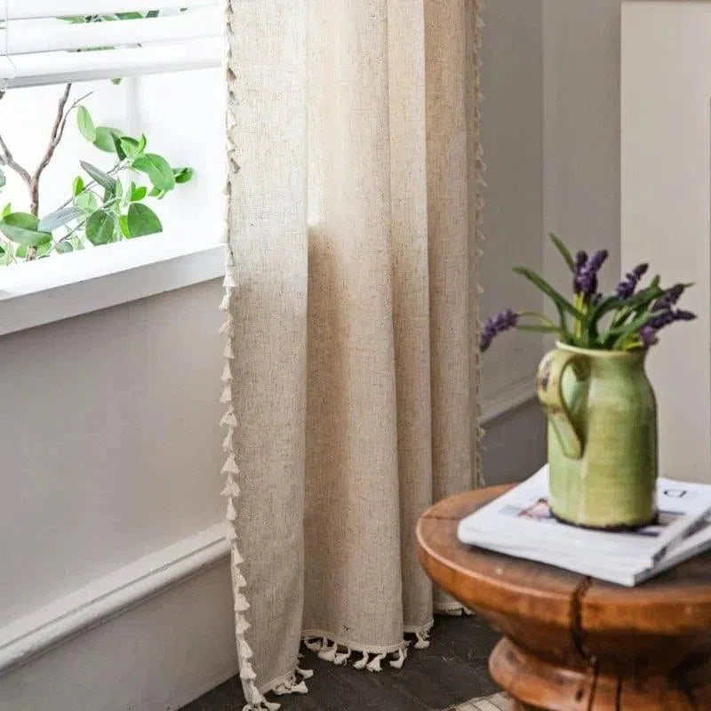Rideau décoratif beige avec bordure à houppes, créant une atmosphère chaleureuse et élégante dans une pièce lumineuse. Accompagné d'une table en bois agrémentée d'un vase vert avec des fleurs violettes, idéal pour un intérieur sophistiqué et naturel. Parfait pour les salons, chambres ou bureaux.